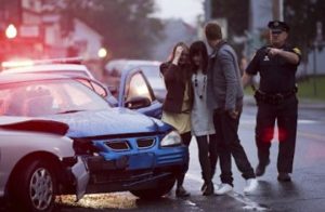 Two women, comforted by a man, stand near a damaged car after an accident as a police officer walks toward them on a wet street.