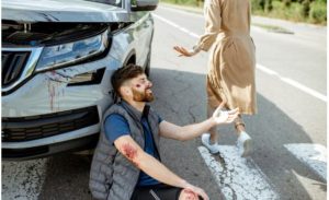 Injured man sitting on the road next to a car with a damaged front, while a person in a trench coat walks away.