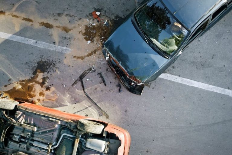 Aerial view of a car accident showing one vehicle flipped over and another car with front-end damage on a street, surrounded by debris and fluid spills.