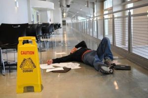 A person lies on the floor near scattered papers and a laptop, next to a yellow caution sign in a hallway.