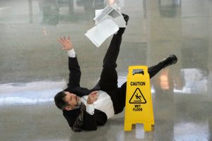 Man in a suit slips near a 'Caution Wet Floor' sign, papers flying in the air, on a shiny floor.