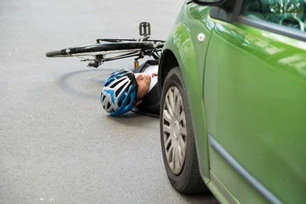A person wearing a blue helmet lies on the ground next to a bicycle and a green car, suggesting a collision or accident.