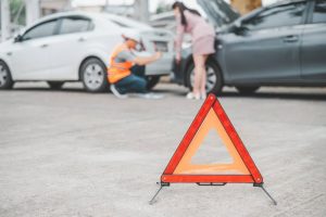 Two cars are stopped on the road with their hoods open. A warning triangle is in the foreground. A person in a safety vest inspects the vehicles while another stands nearby.
