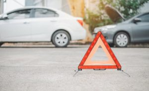 A red caution triangle placed on the road, with two parked cars in the background, one with the hood open.