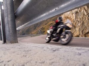 A motorcycle speeds around a curve on a mountain road, captured in motion blur. A metal guardrail is visible in the foreground.