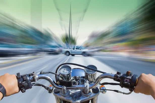 Person riding a motorcycle at high speed on a city street, with blurred surroundings and a white car ahead, suggesting motion.