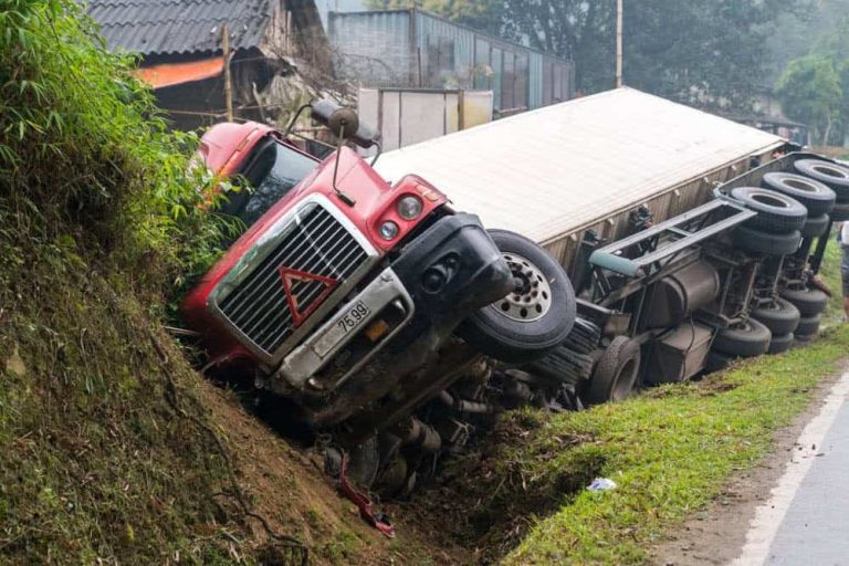 A red semi-truck is overturned on its side in a ditch next to a road, with its cargo trailer partially off the pavement.