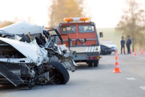 Crashed car on roadside with significant front damage, tow truck and traffic cones nearby, two people in the background.