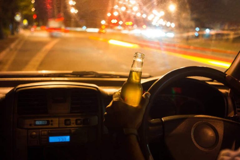Person driving a car at night holding an open beer bottle in their right hand, streetlights and traffic visible through the windshield.