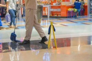 A person in uniform mops the floor of a shopping mall near a yellow caution sign, with stores and shoppers visible in the background.