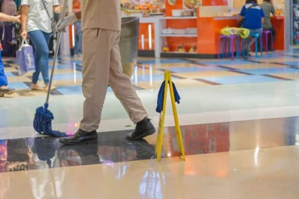 A person in uniform mops the floor of a shopping mall near a yellow caution sign, with stores and shoppers visible in the background.