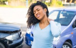 A woman stands between two cars with visible damage, holding her neck and appearing to be in pain, suggesting a recent car accident.