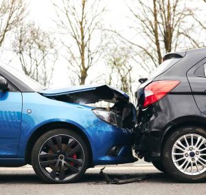 A blue car has collided with the rear of a black car on a road, causing visible damage to both vehicles. Trees without leaves are in the background.