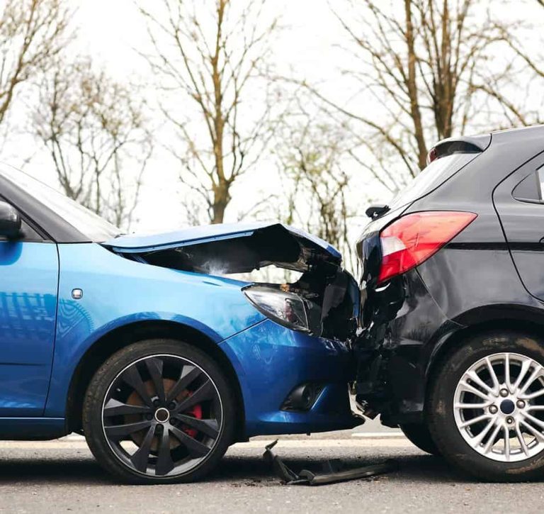 A blue car has collided with the rear of a black car on a road, causing visible damage to both vehicles. Trees without leaves are in the background.