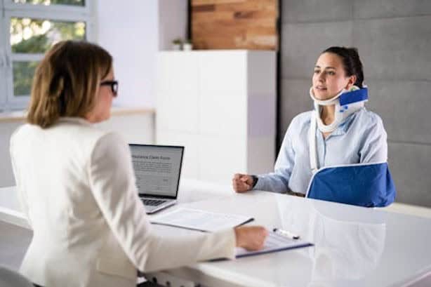A woman wearing a neck brace and arm sling sits across a desk from another woman with a laptop and documents, likely in a professional or medical office setting.