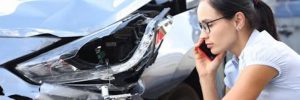 A woman in glasses talks on a phone next to a damaged car headlight.