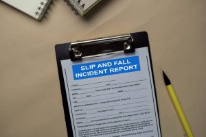 A clipboard with a "Slip and Fall Incident Report" form, next to a yellow pen and two spiral notebooks on a beige surface.