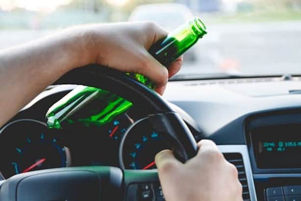 A person holding a green glass bottle and gripping a steering wheel while driving a car.