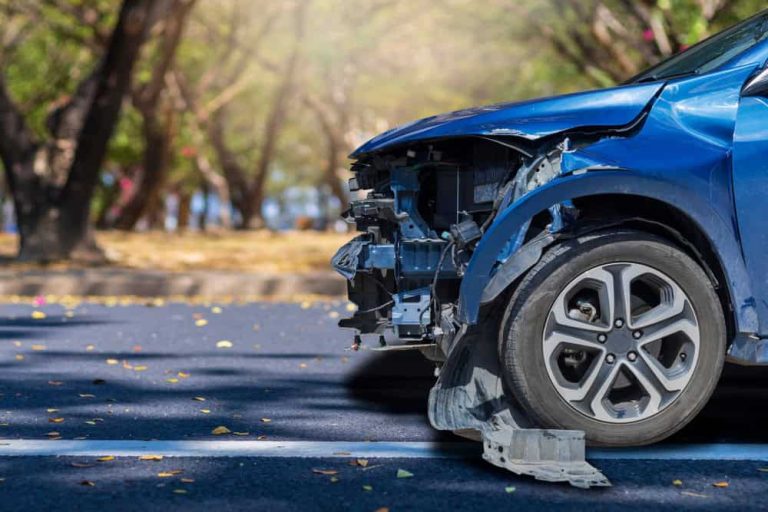 A blue car with a damaged front end and exposed internal parts is parked on a road with trees in the background.