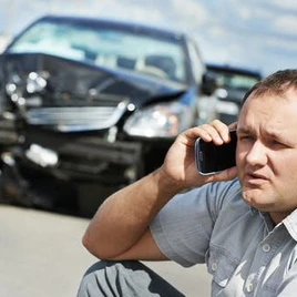 Man talking on phone beside a damaged car after an accident, with another vehicle visible in the background.