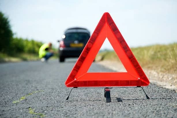A red warning triangle is placed on the road, with a parked car and a person in a high-visibility vest in the background.