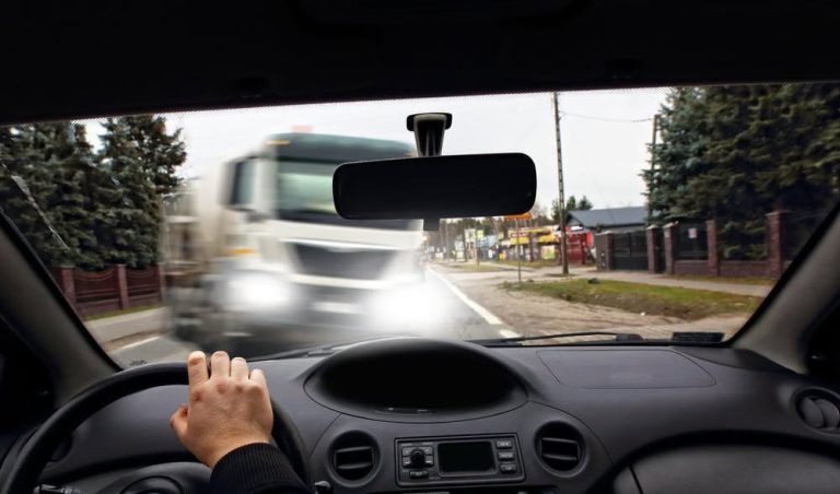 View from inside a car showing a hand on the steering wheel as a large truck approaches quickly from the opposite direction on a two-lane road.