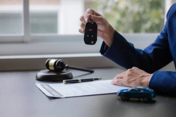 Person in a suit holding a car key over a document on a desk with a gavel, a pen, and a toy car.