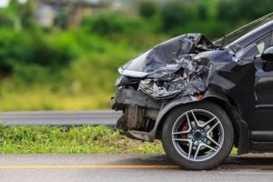 Front view of a black car with significant damage to the front bumper and hood, parked on the side of a road with green foliage in the background.