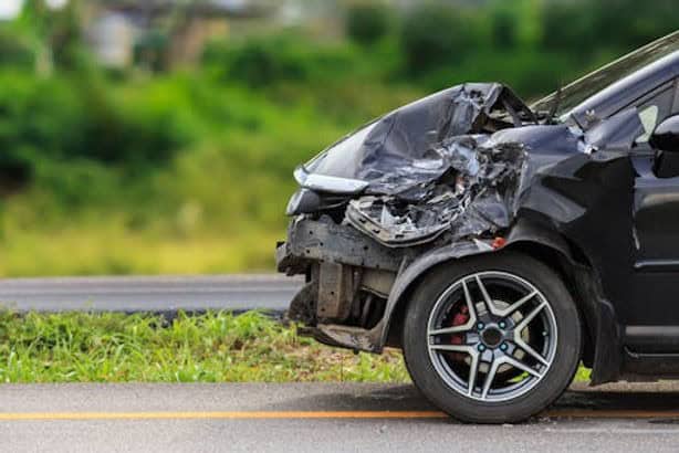 Front view of a black car with significant damage to the front bumper and hood, parked on the side of a road with green foliage in the background.