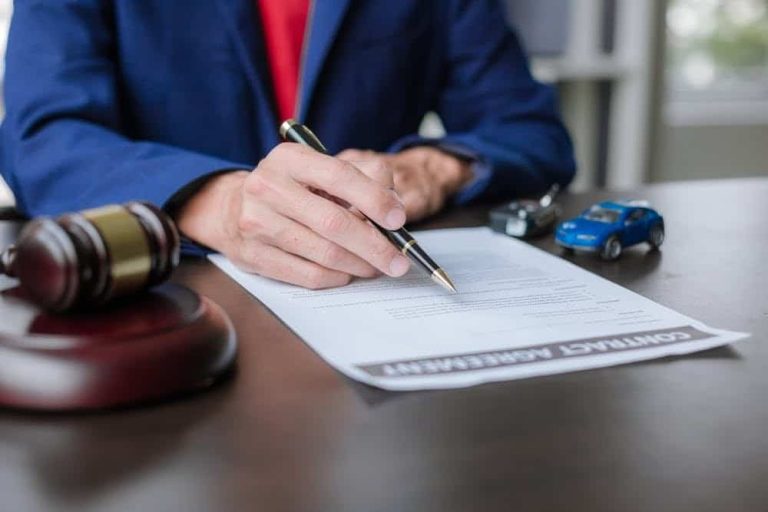 A person signs a contract agreement at a desk with a judge's gavel and two toy cars nearby.