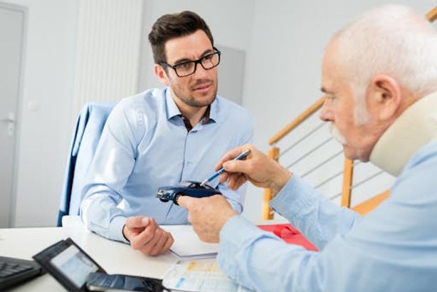 An older man with a neck brace discusses documents and a toy car with a younger man in an office setting.