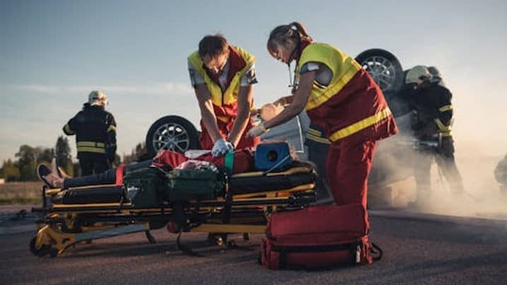 Paramedics attend to an injured person on a stretcher near an overturned car, while firefighters work in the background amid smoke.
