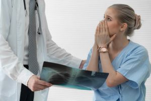 A doctor shows a chest X-ray to a concerned patient, wearing a blue gown, who is covering her mouth with her hands.