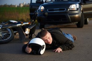 A person lies on the road holding a helmet near a motorcycle and a car, possibly after an accident.