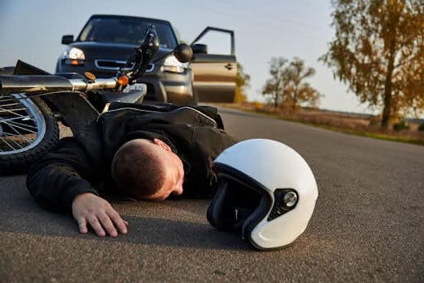 A person lies face down on a road beside a motorcycle and helmet, with a car parked nearby and its driver's door open.