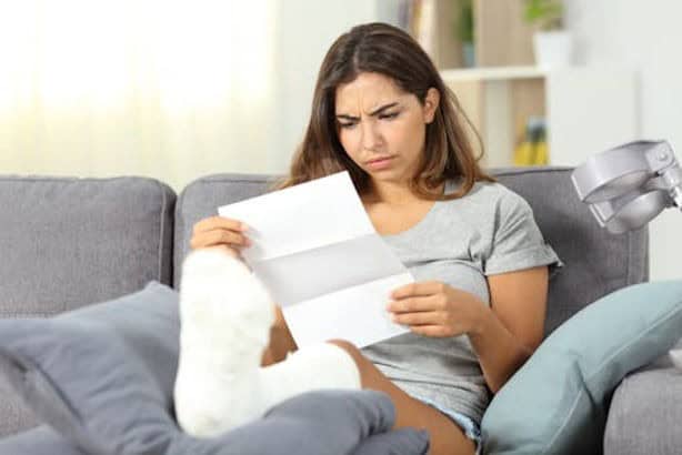 A woman with a bandaged leg sits on a couch, reading a letter with a concerned expression.