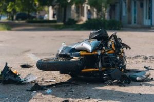 A damaged motorcycle lies on its side on a street, surrounded by debris. Trees and buildings are visible in the background.