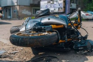 A damaged motorcycle lies on its side on a street, with debris scattered around. A building and parked cars are visible in the background.