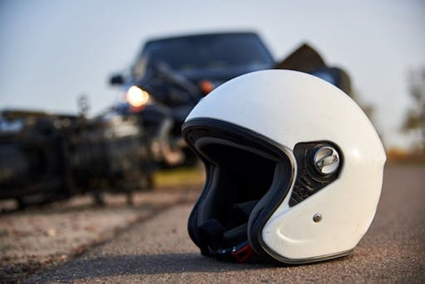 A white motorcycle helmet lies on the road with a blurred motorcycle and car accident scene in the background.