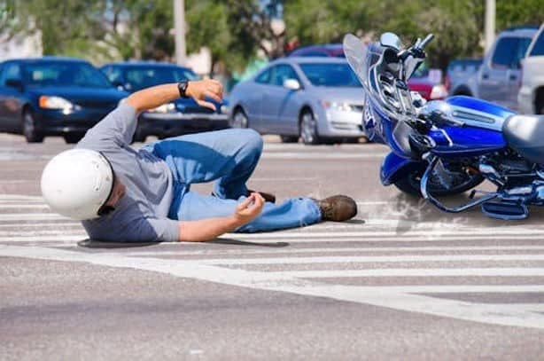 A person wearing a helmet lies on a crosswalk after falling off a blue motorcycle at an intersection, with cars in the background.