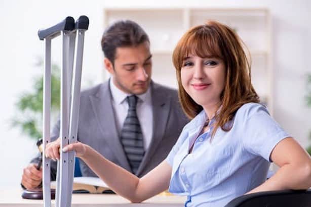 Woman with crutches sits at a desk and smiles at the camera while a man in a suit reads documents in the background.