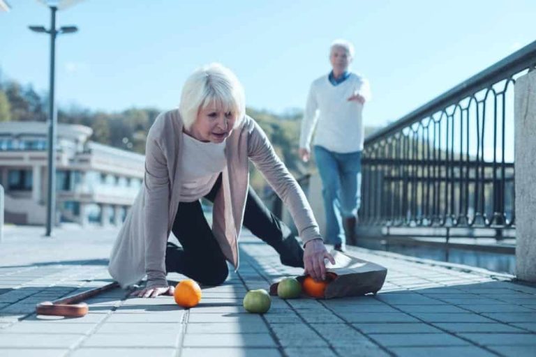 An elderly woman on the ground tries to pick up fallen groceries, while an elderly man approaches and a cane lies nearby.