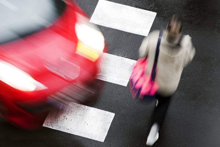 A red car approaches a blurred pedestrian crossing a wet crosswalk, illustrating a potential traffic hazard.
