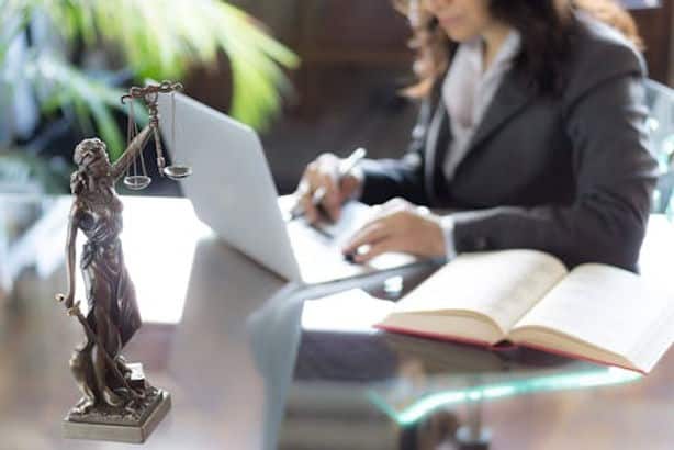 A woman in business attire works on a laptop at a desk with an open book and a bronze Lady Justice statue in the foreground.