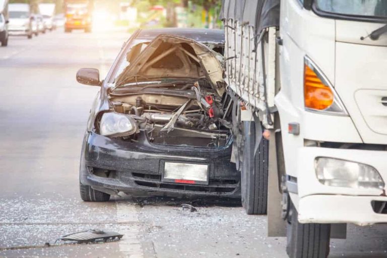 A black car with a heavily damaged front end is crashed into the rear of a white truck on a road, with scattered debris and broken glass visible.