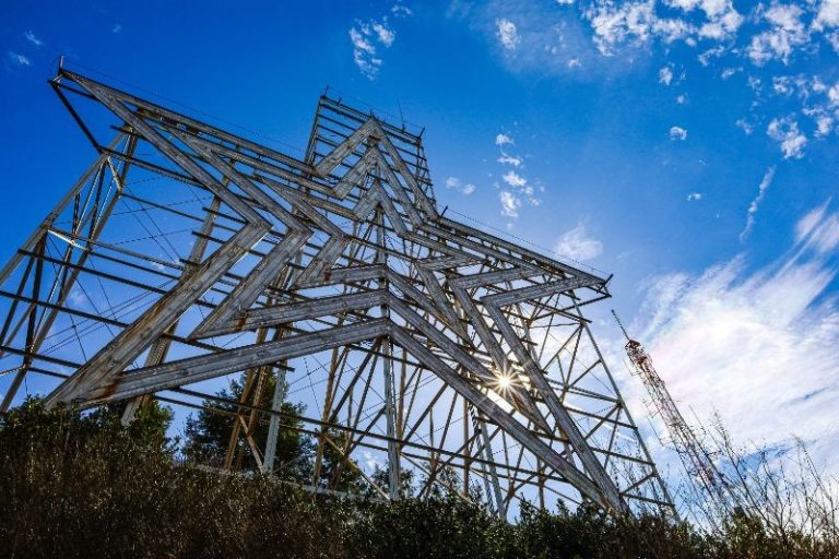 Large metal star structure photographed from below, with sunlight shining through and a blue sky with scattered clouds in the background.