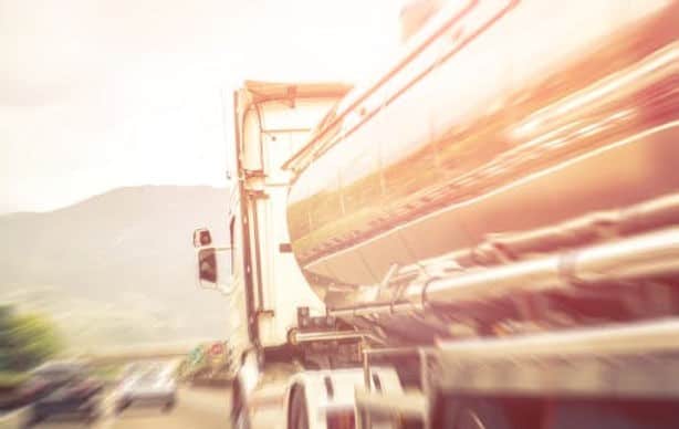 A tanker truck drives on a road with blurred surroundings and distant vehicles, suggesting motion and speed.