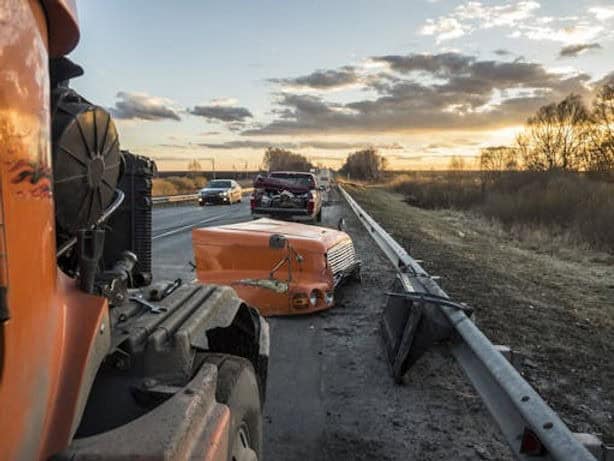 Wrecked vehicles and debris are scattered across a highway after a collision, with damaged car parts next to a guardrail under a cloudy sunset sky.