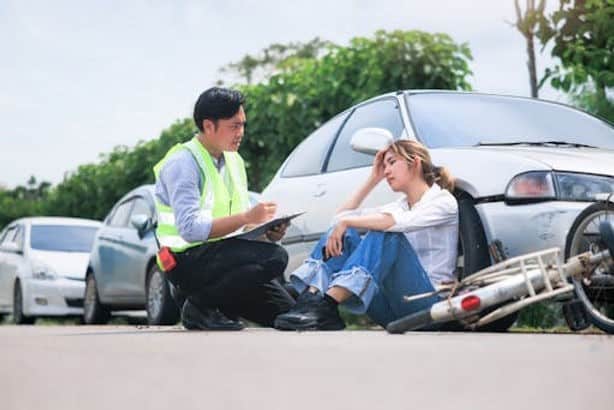 A woman sits on the ground next to a fallen bicycle and a damaged car while a man in a safety vest talks to her and takes notes.