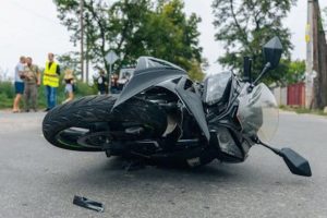 A black motorcycle lies on its side on a road after an accident, with debris scattered nearby and people standing in the background.